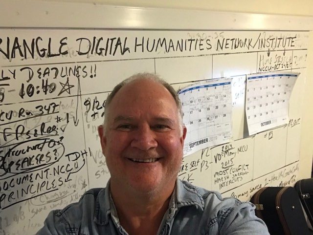 A middle-aged white man smiling in front of a white board covered in notes and schedules.